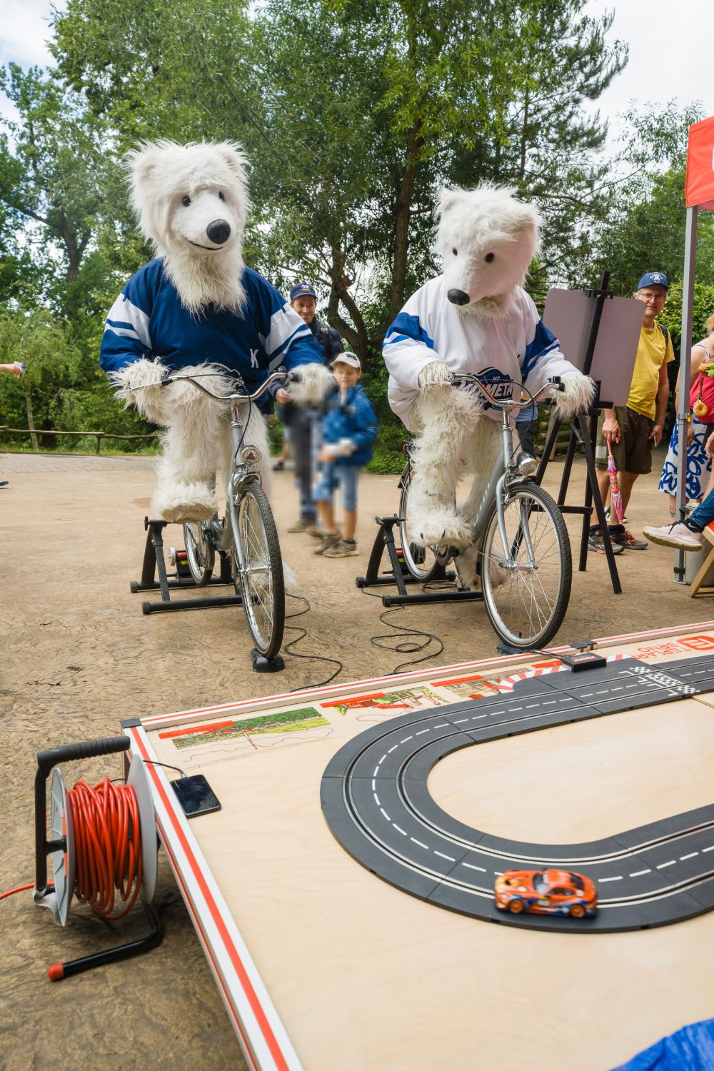Two HC Kometa Brno polar-bear mascots in hockey jerseys pedal bicycles on trainers wired to a slot-car track; an orange toy car races on the track in the foreground.