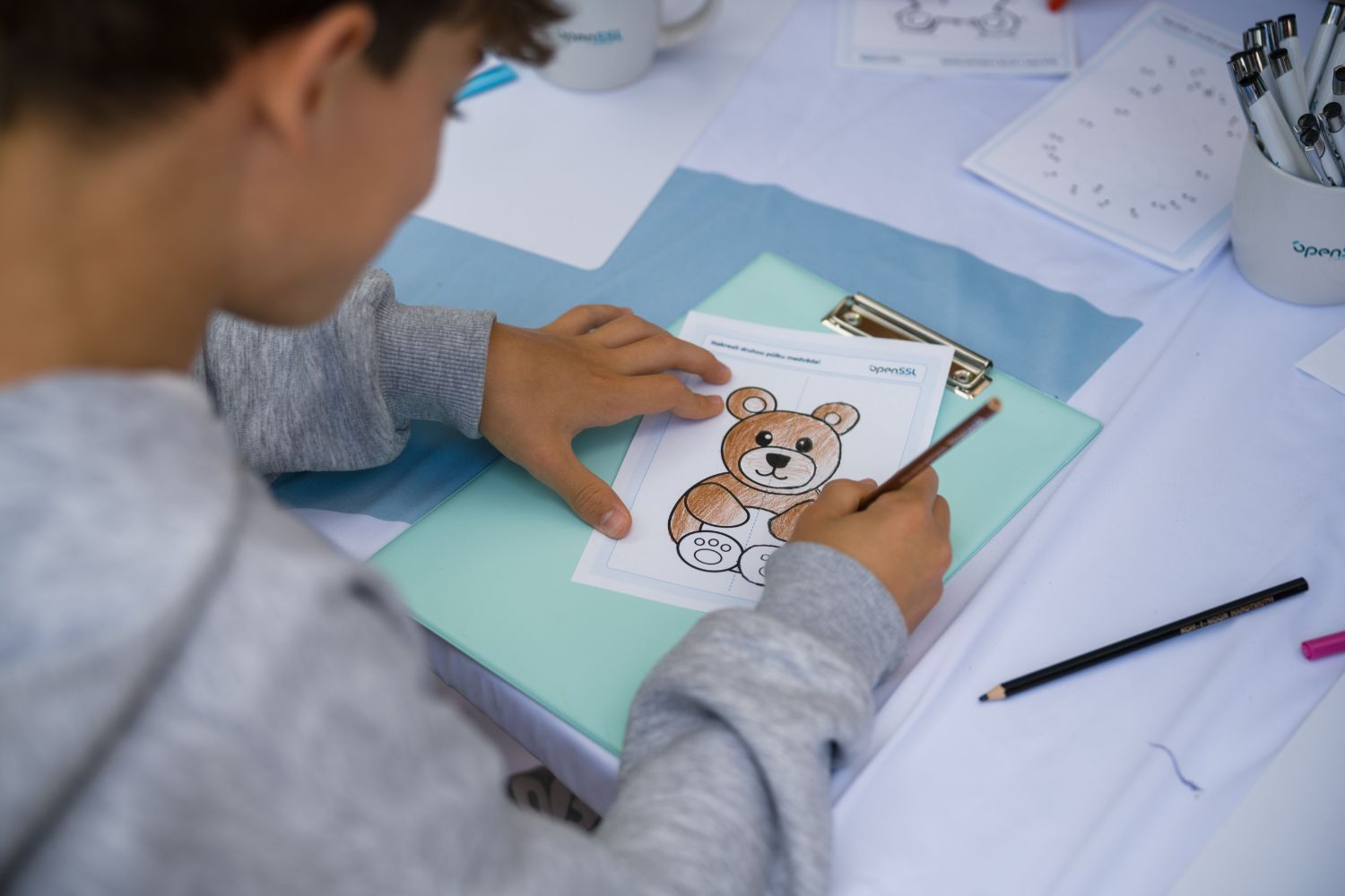 A child leans over the OpenSSL table, carefully colouring a teddy-bear worksheet in brown pencil, an OpenSSL mug of pencils nearby.