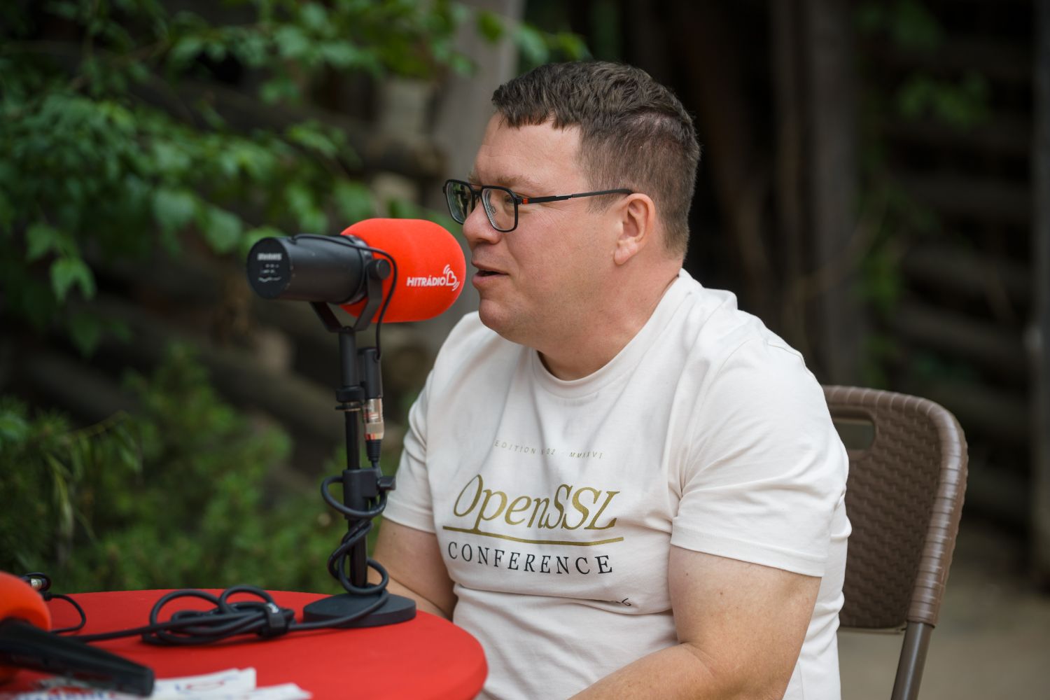 Tomáš, in an OpenSSL Conference t-shirt, speaks into a red Hitrádio microphone during a live broadcast at the zoo, greenery behind him.