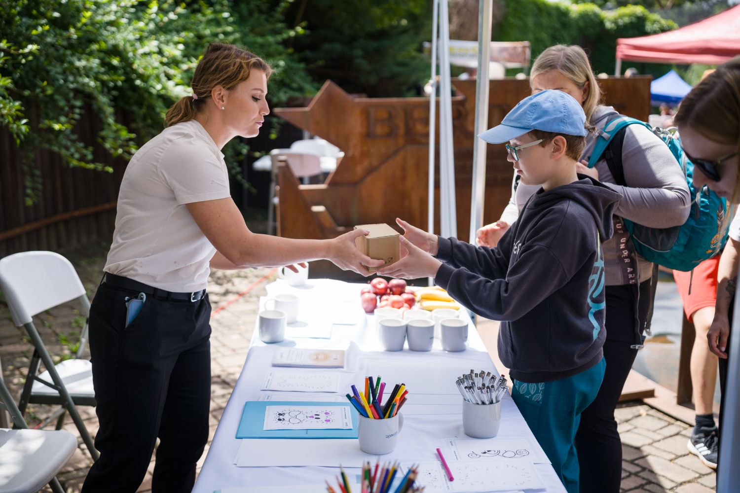 An OpenSSL team member hands a small gift box to a boy across the stand, with apples, bananas and mugs on the table.