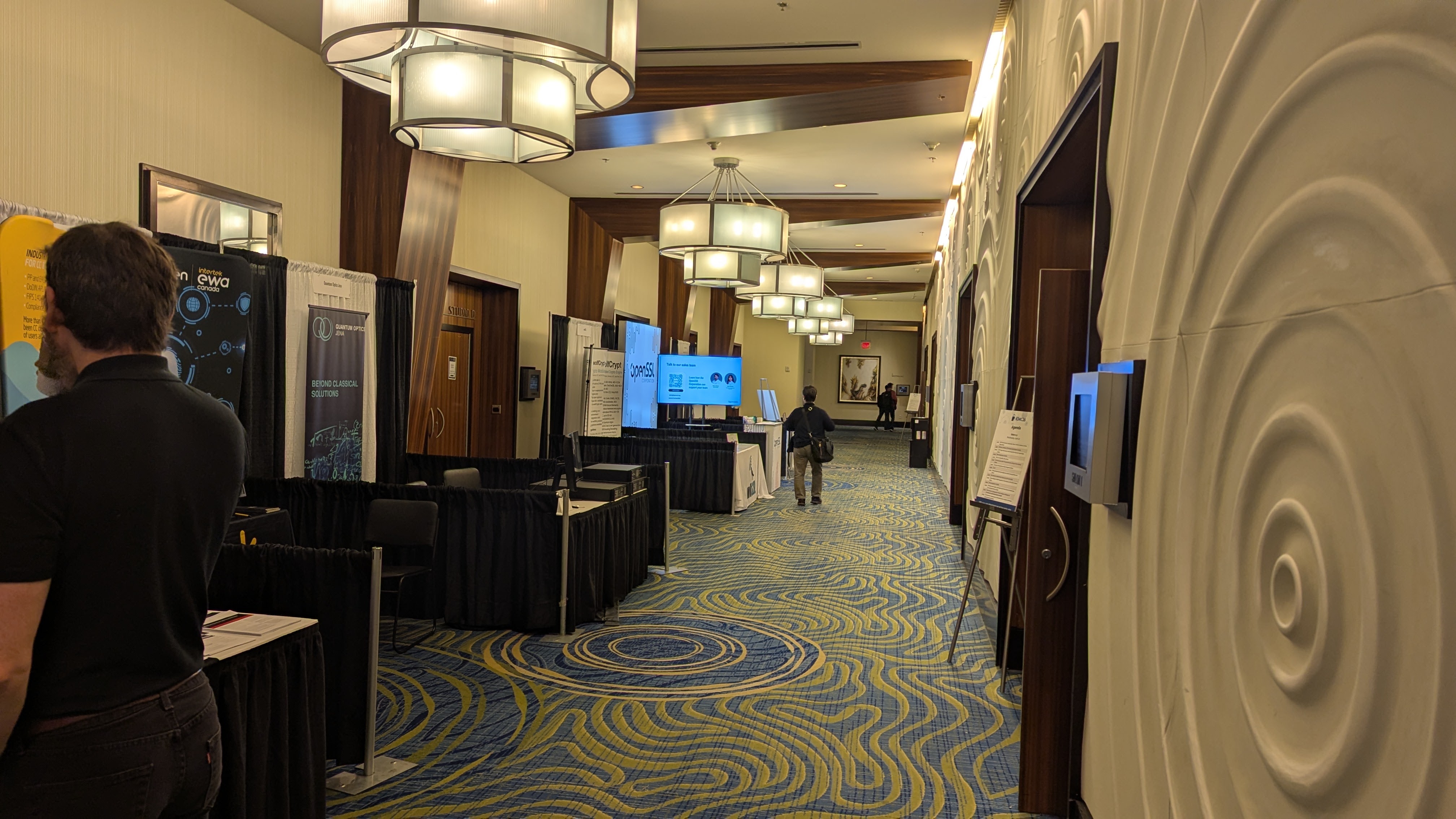 ICMC26 exhibit corridor on Wednesday afternoon, patterned carpet, pendant lights, a single attendee walking.