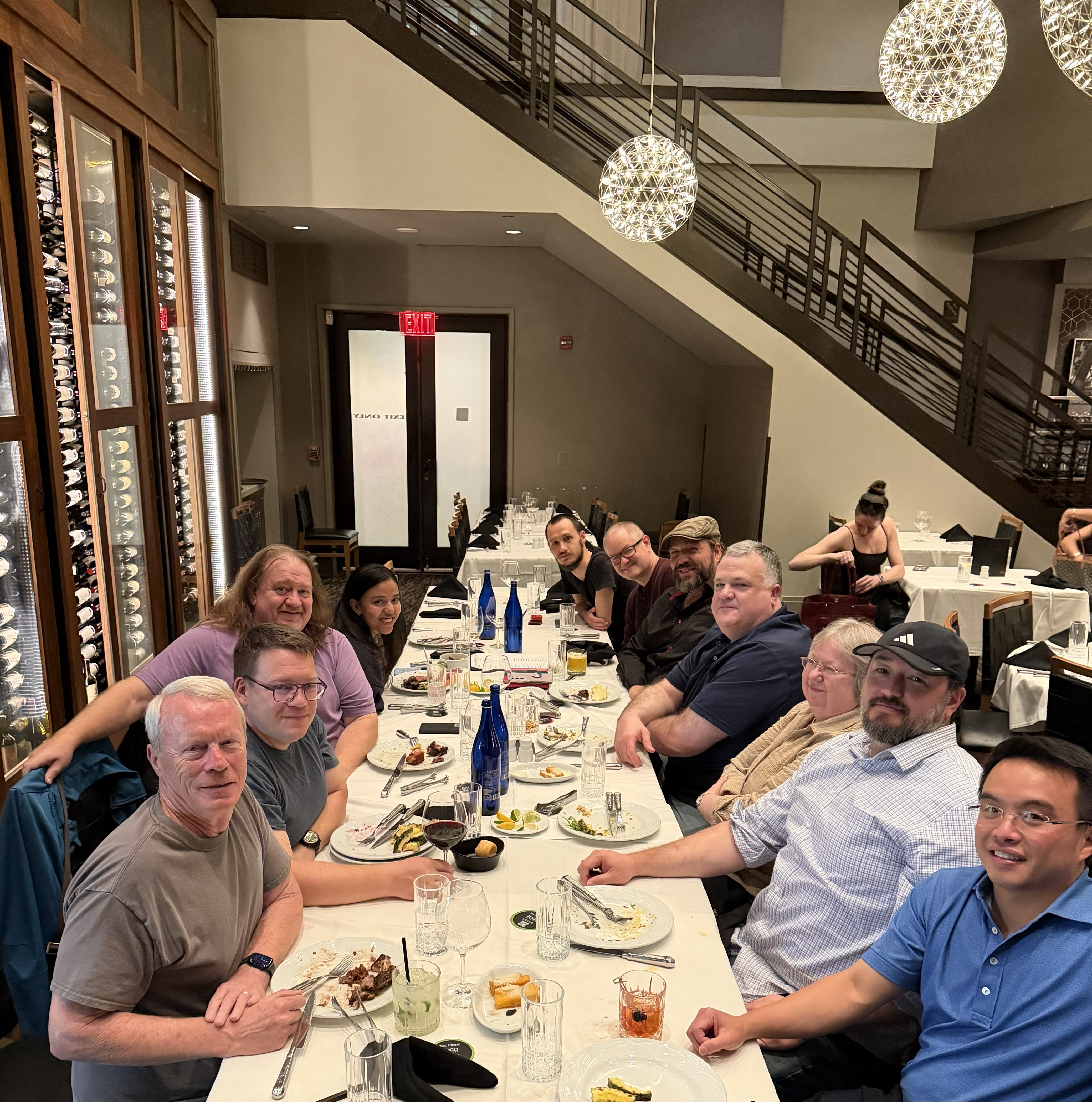 The OpenSSL Corporation team and new friends gathered around a long table at Fogo de Chão, Thursday night of ICMC26 — wine wall to the left, the dining room opening up behind, plates mid-course and glasses in hand.