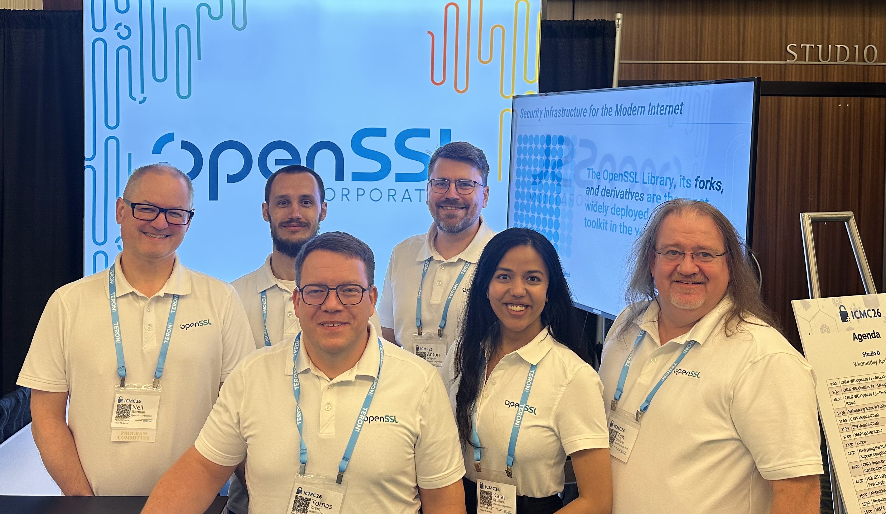 The OpenSSL Corporation team at Booth 4, six members in matching OpenSSL polos, backdrop and display screen behind them.